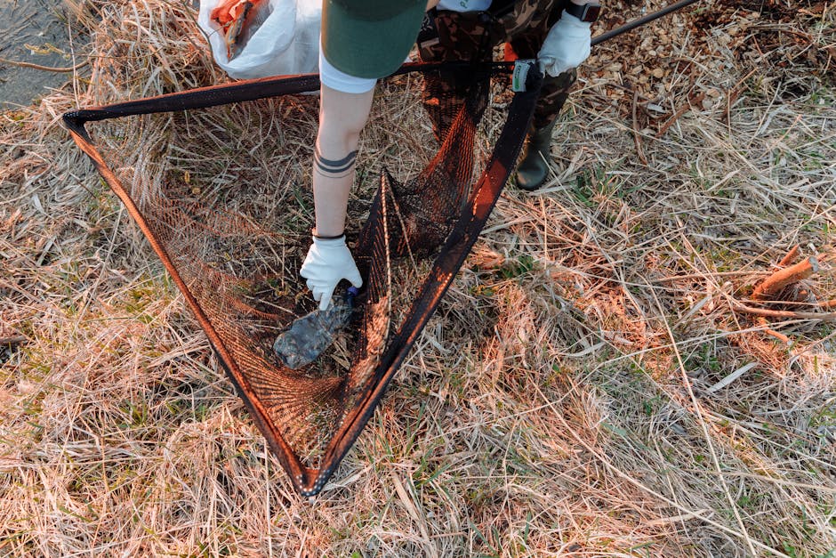 A person wearing gloves, a green cap, and camouflage trousers is kneeling on dry grass and weeds outdoors, collecting a piece of dark, irregularly shaped debris into an orange mesh waste collection ne